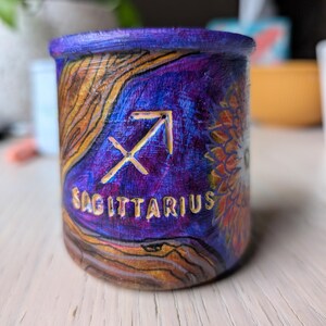 Puede incluir: Un recipiente cilíndrico pintado a mano con el tema de Sagitario. El recipiente presenta una vibrante paleta de colores de azules, púrpuras y dorados, con el símbolo de Sagitario y la palabra "SAGITTARIUS" prominentemente mostrados. El diseño incluye un patrón texturizado similar a la madera.