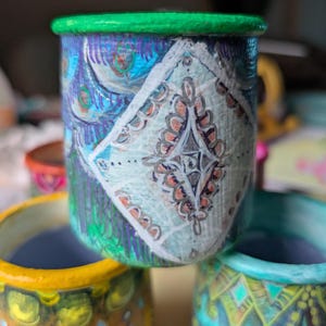 Könnte beinhalten: Ein Stapel aus drei kleinen, handbemalten Keramiktöpfen. Der obere Topf hat einen grünen Rand und ein Design mit blauen, lila und weißen Mustern. Der mittlere Topf hat ein rautenförmiges Design. Die unteren Töpfe sind gelb und türkis.