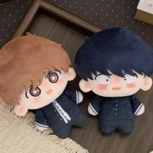 May include: Two plush dolls with large embroidered eyes and soft fabric hair. The left doll has light brown hair and a dark blue suit with white accents. The right doll has dark blue hair, a serious expression, and a matching dark blue suit.