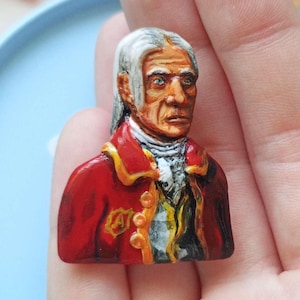May include: A small, hand-painted pin featuring a bust of an older figure with white hair, wearing a red coat with gold trim and a white cravat. The figure has a weathered face with visible wrinkles and a gold letter A emblem on the chest.