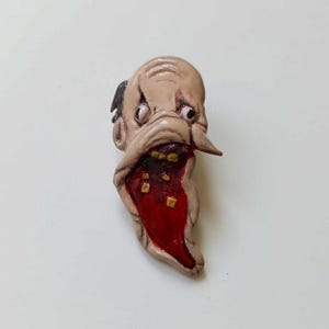 May include: A sculpted pin featuring a pale, distorted human-like face with a wide, gaping mouth. The interior of the mouth is painted a deep, glossy red with small, scattered yellow square accents. The eyes are wide and dark, with skin folds.