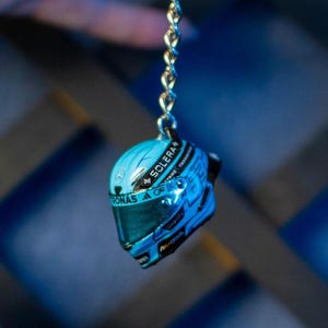 May include: A miniature blue and black racing helmet keyring. The keyring is suspended by a silver chain. The helmet has the word "SOLERA" printed on it. The background is a blurred blue and black.