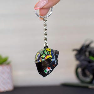 Può includere: Un portachiavi a forma di casco da moto colorato con visiera nera e logo AGV. Il portachiavi è attaccato a una catena argentata.