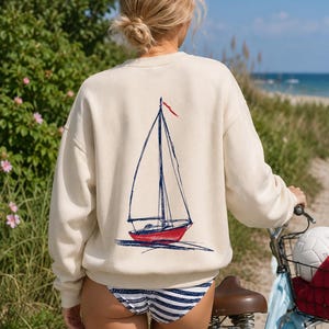 Peut inclure: Un sweat-shirt à manches longues de couleur crème avec une illustration graphique d'un voilier au dos. Le voilier possède une coque rouge, un mât et un gréement bleu foncé, ainsi qu'un petit drapeau rouge au sommet. Col rond.