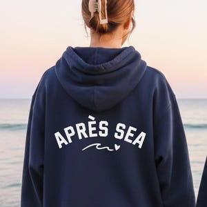 Może przedstawiać: Dwie granatowe bluzy z kapturem z białym napisem "APRÈS SEA" nad białym wzorem fali. Bluzy z kapturem są noszone przez dwie osoby stojące na plaży, zwrócone w stronę morza.
