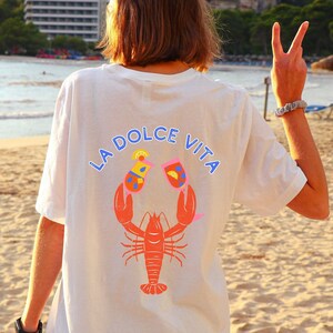 May include: White t-shirt with a graphic design on the back. The design features a red lobster holding two cocktails, with the words "LA DOLCE VITA" in blue above. The shirt is worn on a beach.