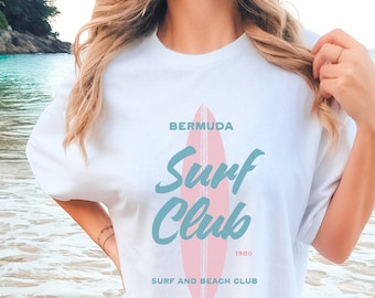 Könnte beinhalten: Ein weißes T-Shirt mit einer blauen und pinkfarbenen Grafik eines Surfboards und dem Text "Bermuda Surf Club 1980 Surf and Beach Club". Die Person, die das Shirt trägt, steht im Meer.