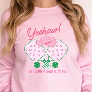 Op de afbeelding: Roze sweatshirt met een afbeelding van twee gekruiste pickleball-rackets met een roze cowboyhoed erop. De tekst "Yeehaw! Let's Pickleball, Y'all" staat op de sweatshirt gedrukt.