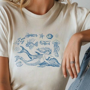 Puede incluir: Una camiseta blanca con un gráfico azul de una sirena montando un delfín rodeada de otras criaturas marinas, incluyendo peces, una estrella de mar y una medusa.