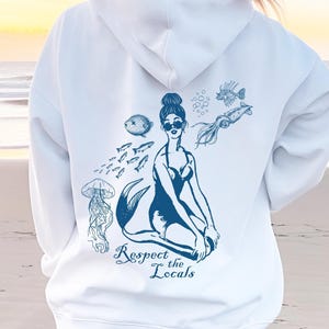 Puede incluir: Sudadera blanca con un estampado gráfico azul de una sirena con gafas de sol, rodeada de peces y burbujas. El texto "Respect the Locals" está impreso debajo de la sirena.