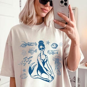Puede incluir: Camiseta beige de gran tamaño con una ilustración azul de una sirena y criaturas marinas. El diseño incluye una sirena con gafas de sol, peces y una medusa. La modelo lleva un gorro amarillo y gafas de sol negras.