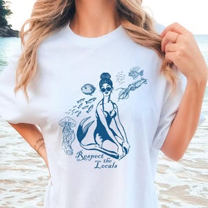 Puede incluir: Camiseta blanca con un gráfico azul de una sirena con gafas de sol y el texto "Respect the Locals".