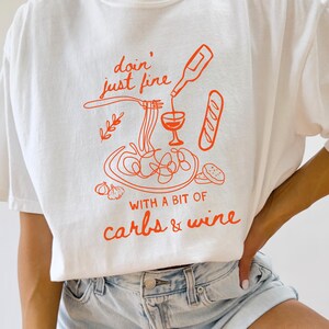 May include: White t-shirt with an orange graphic of pasta, wine, and bread. The text on the shirt reads "doin' just fine with a bit of carbs & wine". The shirt is a casual, comfortable style.