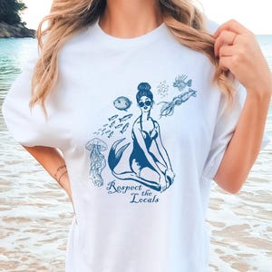 Puede incluir: Camiseta blanca con un gráfico azul de una sirena con gafas de sol, rodeada de peces, una medusa y el texto "Respect the Locals".