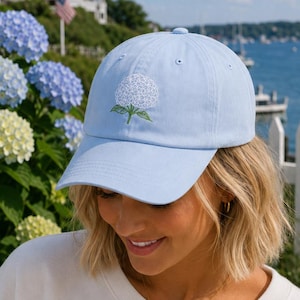 Puede incluir: Una gorra de béisbol de color azul claro con una flor de hortensia blanca bordada con hojas verdes en el panel frontal. La gorra tiene una visera curva y una corona estructurada, confeccionada en tela sólida con costuras a juego.