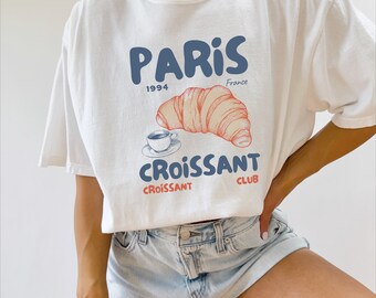 Puede incluir: Camiseta blanca con un diseño gráfico azul y rojo que presenta un croissant, una taza de café y el texto "Paris 1994 France Croissant Club".