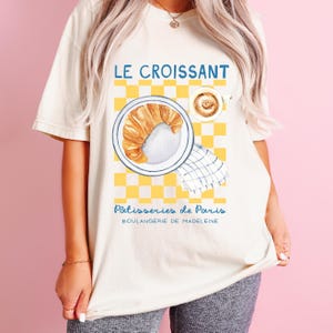 Puede incluir: Camiseta color crema con una ilustraci&oacute;n en acuarela de un cruas&aacute;n en un plato, una taza de caf&eacute; y un fondo a cuadros amarillos. El texto "LE CROISSANT" y "P&acirc;tisseries de Paris" est&aacute;n impresos en la camiseta.