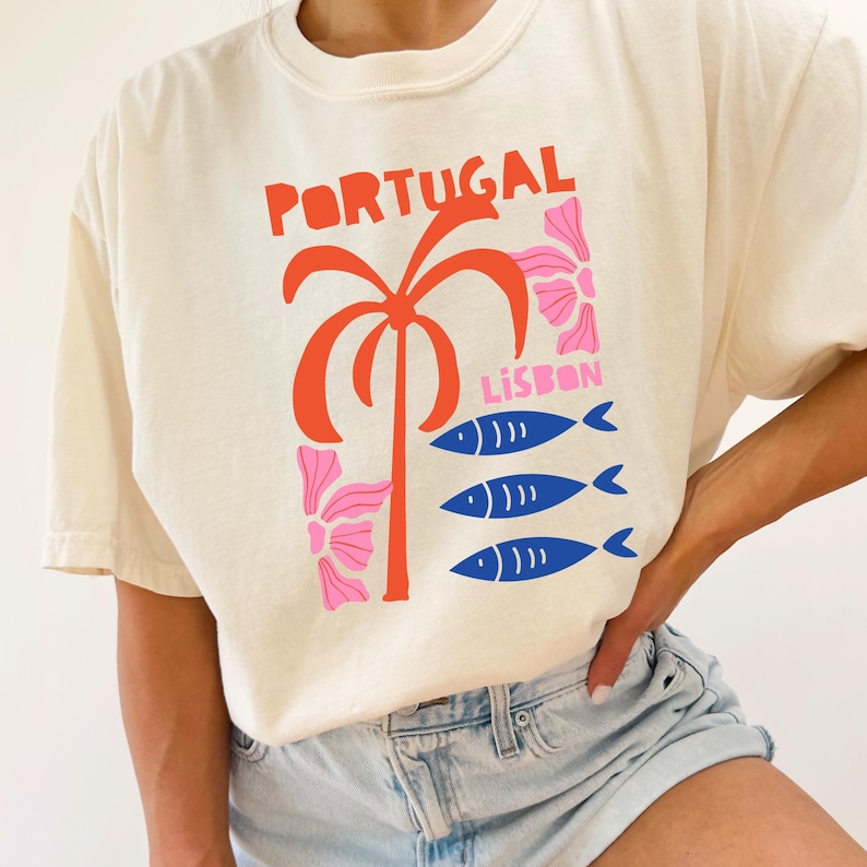 Peut inclure: T-shirt cr&egrave;me avec un motif graphique color&eacute;. Le motif comprend le mot "PORTUGAL" en rouge, un palmier orange stylis&eacute;, des &eacute;l&eacute;ments floraux roses et trois poissons bleus avec le mot "LISBON".