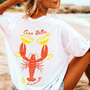 May include: White t-shirt with a red lobster holding two yellow glasses with lemon slices. The text "Ciao Bella Limoncello" is printed on the shirt.