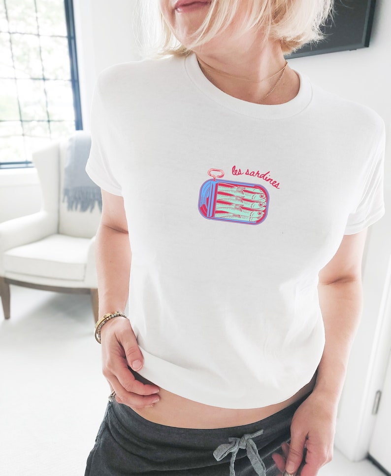 May include: White t-shirt with a graphic of a can of sardines with the text "les sardines" on it.