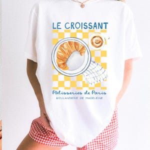 Puede incluir: Camiseta blanca con una ilustraci&oacute;n de acuarela de un cruas&aacute;n en un plato, una taza de caf&eacute; y un patr&oacute;n de cuadros amarillos y blancos. La camiseta tiene el texto "LE CROISSANT" y "P&acirc;tisseries de Paris, Boulangerie de Madeleine."