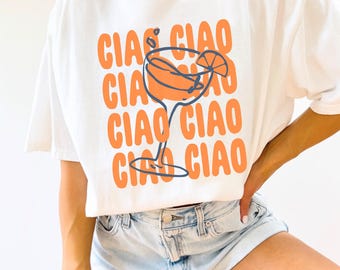 May include: White t-shirt with the text "CIAO CIAO CIAO CIAO CIAO" in orange letters and a stylized orange cocktail glass with a lemon slice on top.