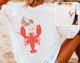 May include: White t-shirt featuring a red lobster graphic with a cocktail and the words "ciao bella". The design is on the back of the shirt, with a smaller lobster graphic on the front. The shirt is a casual, beach-themed design.
