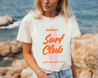 Könnte beinhalten: Weißes T-Shirt mit einer pinkfarbenen und orangen Grafik eines Surfbretts und dem Text "Monaco Surf Club 1980 Surf and Beach Club".