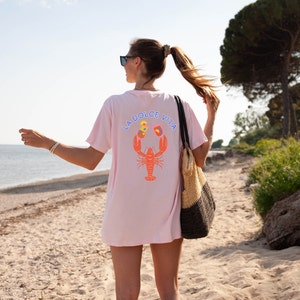 May include: A light pink t-shirt with the words "LA DOLCE VITA" above a lobster graphic. The lobster is orange with colorful accents. The shirt is worn on a beach with a straw tote bag.