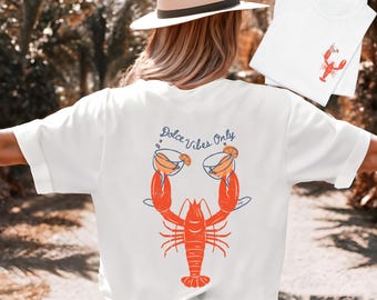 May include: White t-shirt featuring a red lobster design with two cocktails and the text "Dolce Vibes Only." A smaller white t-shirt with a lobster graphic is also visible. The design is in orange and blue.