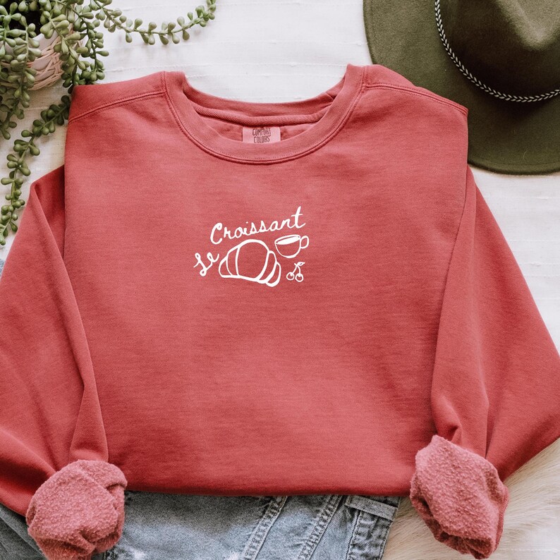 May include: A red sweatshirt with a white graphic design that says "Le Croissant" with a croissant and a cup of coffee.