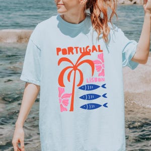 Peut inclure: T-shirt bleu clair avec un motif graphique. Le motif comprend le mot "PORTUGAL" en rouge, un palmier rouge, des fleurs roses, le mot "LISBON" et trois poissons bleus.