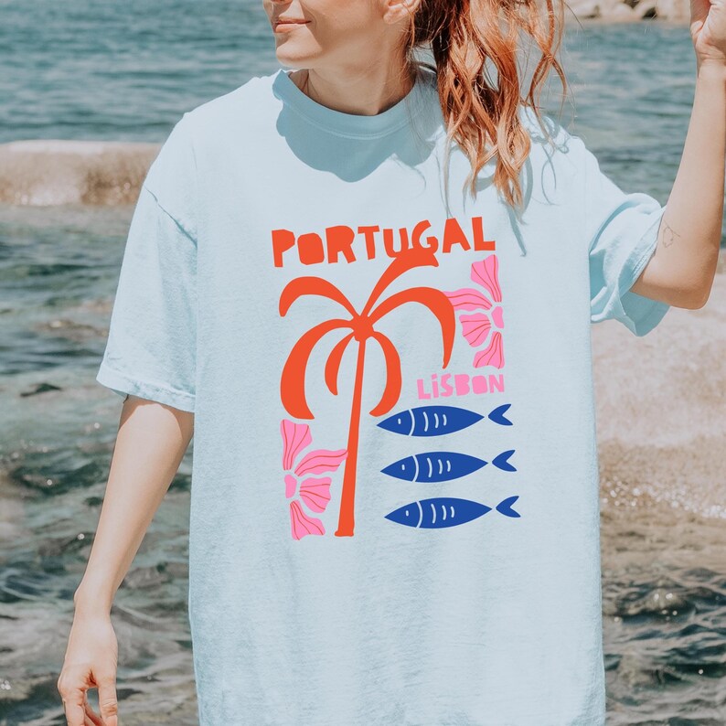 Peut inclure: T-shirt bleu clair avec un motif graphique. Le motif comprend le mot "PORTUGAL" en rouge, un palmier rouge, des fleurs roses, le mot "LISBON" et trois poissons bleus.