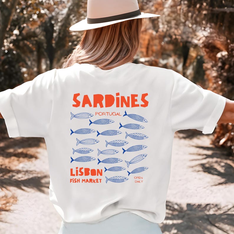 Peut inclure: T-shirt blanc avec le mot "Sardines" en orange, surmontant des illustrations de poissons bleus. Le texte "Lisbon Fish Market" est &eacute;galement pr&eacute;sent. Le design s'inspire de la culture portugaise.