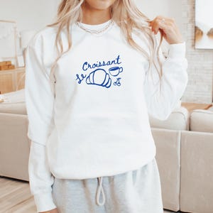 May include: White sweatshirt with blue text that says "Le Croissant" with a drawing of a croissant, a cup of coffee, and two cherries.