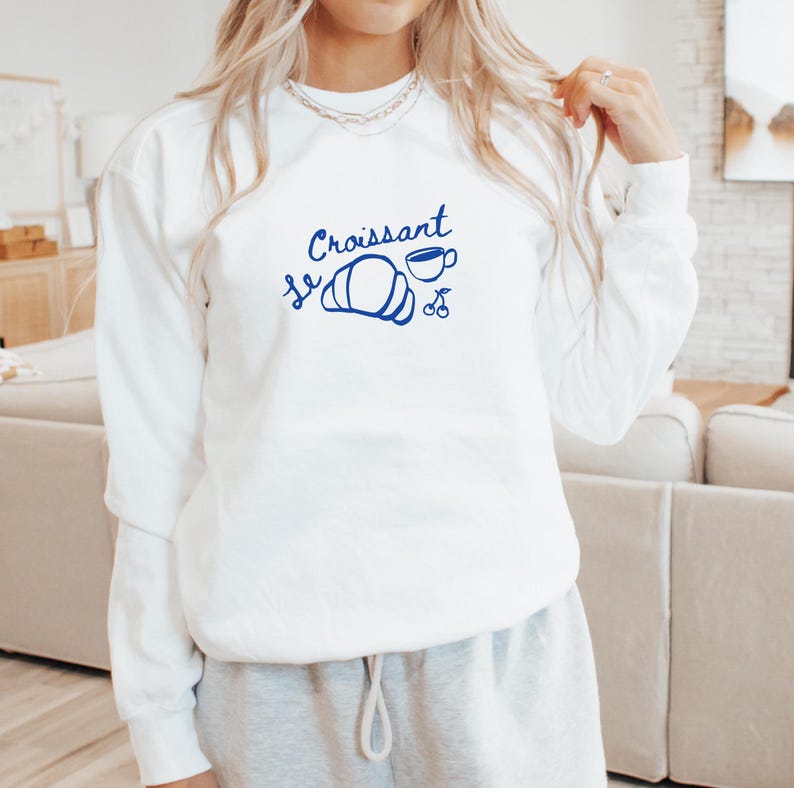 May include: White sweatshirt with blue text that says "Le Croissant" with a drawing of a croissant, a cup of coffee, and two cherries.