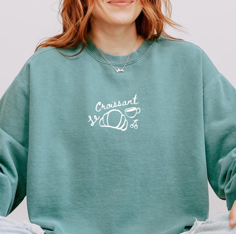 May include: A teal blue sweatshirt with a white graphic design that says "Le Croissant" with a croissant, a cup of coffee, and two cherries.