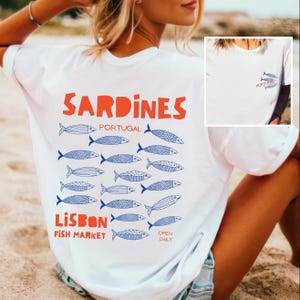 Peut inclure: T-shirt blanc avec un motif orange "Sardines", des illustrations de poissons bleus et le texte bleu "Lisbon Fish Market". Le t-shirt pr&eacute;sente &eacute;galement un petit motif avant avec deux poissons et le mot "Portugal".