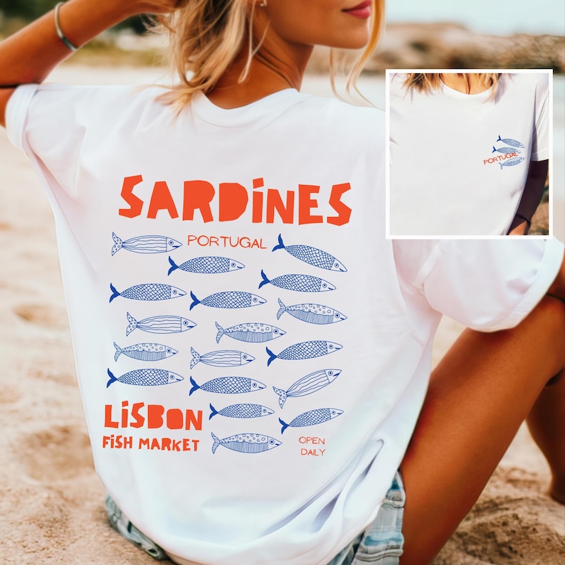 Peut inclure: T-shirt blanc avec un motif orange "Sardines", des illustrations de poissons bleus et le texte bleu "Lisbon Fish Market". Le t-shirt pr&eacute;sente &eacute;galement un petit motif avant avec deux poissons et le mot "Portugal".
