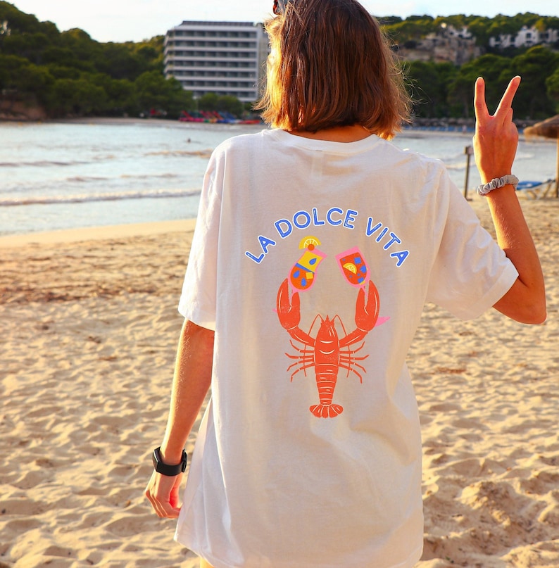 May include: White t-shirt with a graphic design on the back. The design features a red lobster holding two cocktails, with the words "LA DOLCE VITA" in blue above. The shirt is worn on a beach.