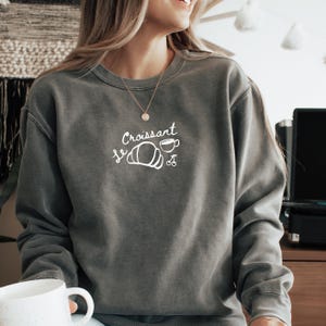 May include: A gray sweatshirt with the text "Le Croissant" in white, featuring a white illustration of a croissant and a cup of coffee.