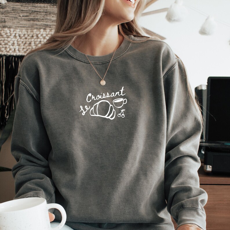 May include: A gray sweatshirt with the text "Le Croissant" in white, featuring a white illustration of a croissant and a cup of coffee.