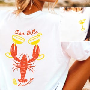 May include: White t-shirt with a red lobster holding two yellow glasses of limoncello. The text "Ciao Bella" is above the lobster and "Limoncello" is below.