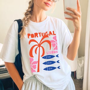 Peut inclure: T-shirt blanc avec un motif graphique color&eacute; repr&eacute;sentant le mot "PORTUGAL" en orange, un palmier, des fleurs roses et le mot "LISBON" avec trois poissons bleus. Le t-shirt est de style d&eacute;contract&eacute;.