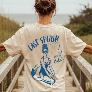 May include: A white t-shirt with a blue graphic of a mermaid with the text "Last Splash" and "She's the Catch" on the back.
