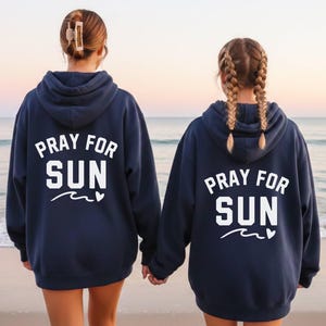Op de afbeelding: Twee marineblauwe hoodies met de witte tekst "PRAY FOR SUN" en een golfafbeelding. De hoodies worden gedragen door twee mensen op een strand, met de oceaan op de achtergrond.