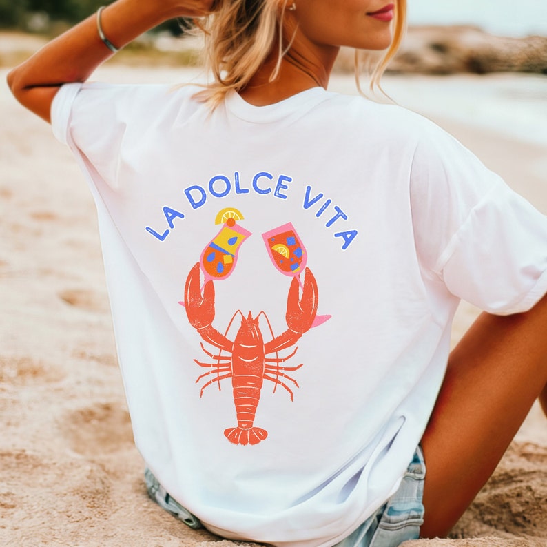 May include: White t-shirt with a graphic design on the back. The design features a red lobster holding two cocktail glasses, with the words "LA DOLCE VITA" in blue above. The cocktails have orange, yellow, and blue details.