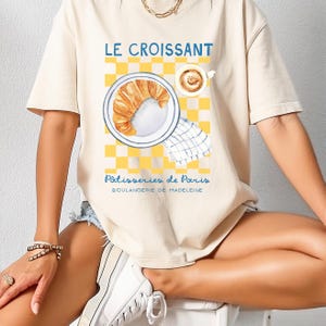 Puede incluir: Camiseta beige con una ilustraci&oacute;n de acuarela de un cruas&aacute;n en un plato, una taza de caf&eacute; y un fondo a cuadros amarillos y blancos. El texto "LE CROISSANT" y "P&acirc;tisseries de Paris" est&aacute; impreso.