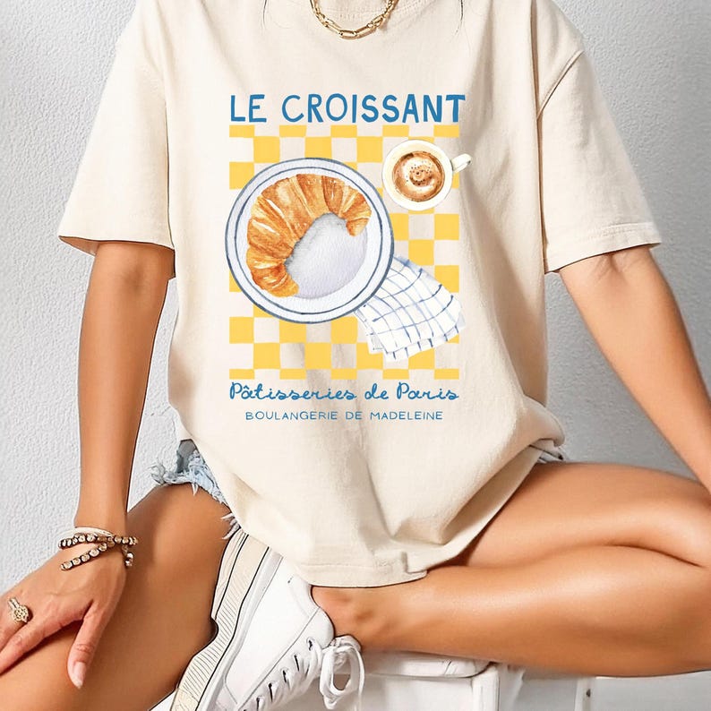 Puede incluir: Camiseta beige con una ilustraci&oacute;n de acuarela de un cruas&aacute;n en un plato, una taza de caf&eacute; y un fondo a cuadros amarillos y blancos. El texto "LE CROISSANT" y "P&acirc;tisseries de Paris" est&aacute; impreso.