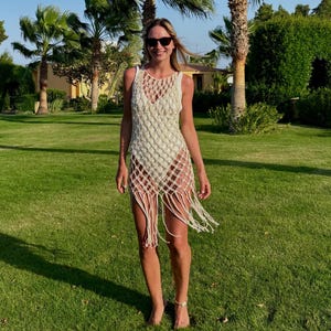 May include: A cream-colored, sleeveless macrame dress with fringe detailing. The dress has a diamond-shaped net pattern and is worn outdoors on a sunny day. The background features green grass, palm trees, and a clear blue sky.
